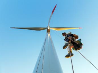 Låg bildvinkel på reparatör och industriklättrare som hänger i lina på vindkraftverk belyst av solnedgången klädd i skoskydd och tittar in i kameran.